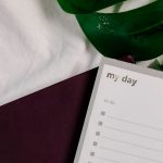 A close-up of a to-do list notebook with lush green leaves on a white textile surface.